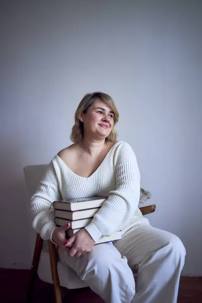 medium-sized woman in light clothes reads a book while sitting in a white chair in a light room