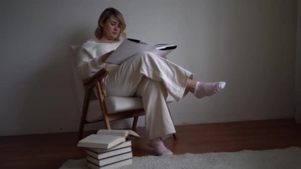 穿着浅色衣服的中等身材女人坐在明亮房间的白色椅子上看书 — 图库视频影像