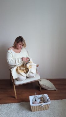 Açık renk elbiseli orta boylu bir kadın, beyaz bir odada beyaz bir sandalyede otururken altın ve bej renkte bir resim nakışlıyor.
