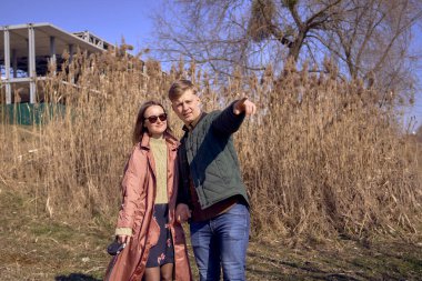 Genç ve şık bir çift nehir kenarında köpekleriyle yürüyor. Arka planda bitmemiş bir park yeri var.            