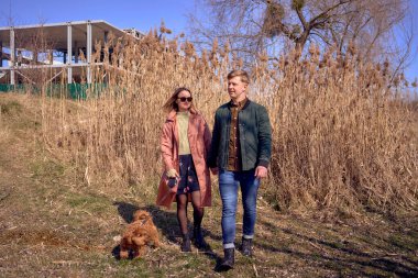 Genç ve şık bir çift nehir kenarında köpekleriyle yürüyor. Arka planda bitmemiş bir park yeri var.            