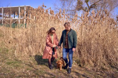 Genç ve şık bir çift nehir kenarında köpekleriyle yürüyor. Arka planda bitmemiş bir park yeri var.            