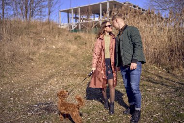 Genç ve şık bir çift nehir kenarında köpekleriyle yürüyor. Arka planda bitmemiş bir park yeri var.            
