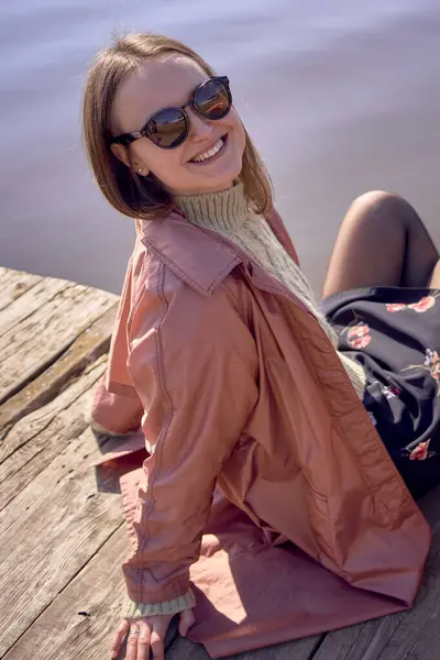 戴着太阳镜的女孩坐在码头上 对着相机微笑 — 图库照片