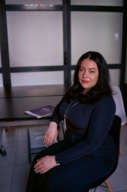                  Orta boylu, siyah saçlı, ofiste siyah elbiseli bir kadının portresi.              