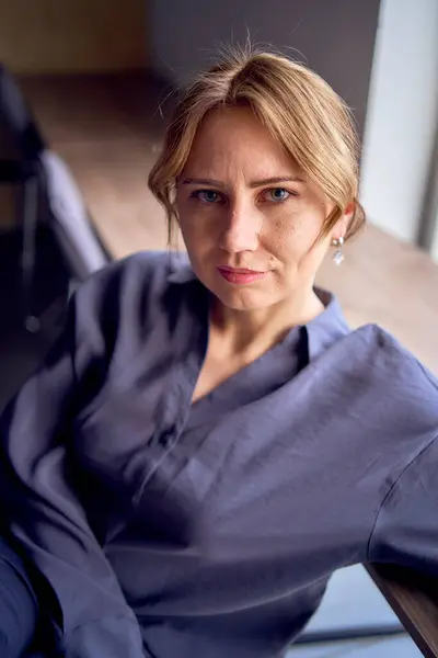 a woman sitting on chair with  legs crossed over the armrest. High heels, black pants, silk blouse. Cityscape outside through window. Modern interior design with neutral tones.