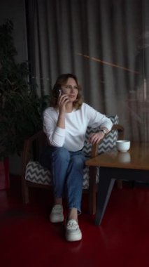  Kazaklı ve kotlu bir kadın, arka planda yeşil paneller olan güzel bir kafede telefonla konuşuyor.                    