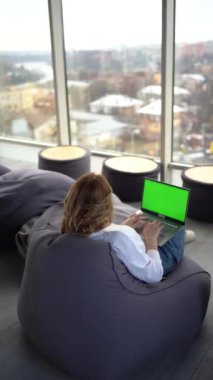 Günlük kıyafetli bir kadın panoramik pencerenin önündeki bir laptopun üzerinde oturuyor. Şehir manzarası, yeşil ekran, Chroma anahtarı.