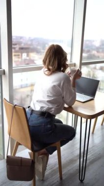 Kazaklı ve kot pantolonlu bir kadın kahve içer ve panoramik pencereli ve şehir manzaralı bir çalışma alanında dizüstü bilgisayarda çalışır.       
