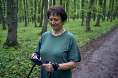  an elderly woman doing Nordic walking with sticks in spring forest switches podcast on phone              clipart