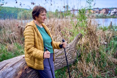 Yaşlı bir kadın, İskandinav yürüyüş egzersizinden sonra nehir kıyısında dinleniyor