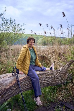 Yaşlı bir kadın, İskandinav yürüyüş egzersizinden sonra nehir kıyısında dinleniyor