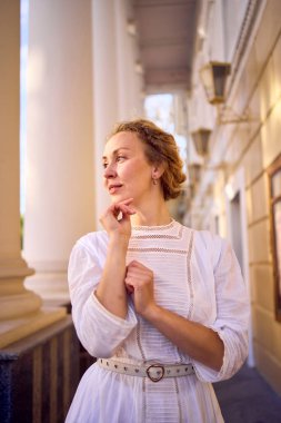 Tiyatronun yanında antika sütunları olan, beyaz, klasik, zarif bir orta yaşlı kadın.
