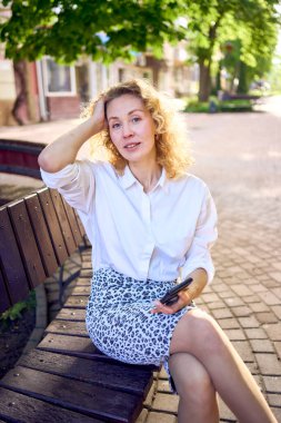  70 'li yaşlarda güzel, orta yaşlı bir kadın, 80' li yaşlarda bir elbise, akıllı telefonuna bir bankta bakıyor.