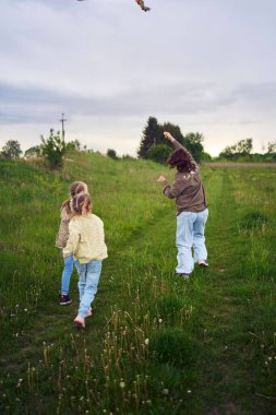 İki küçük kız kardeş ve anne koşup tarlaya uçurtma fırlatıyorlar.