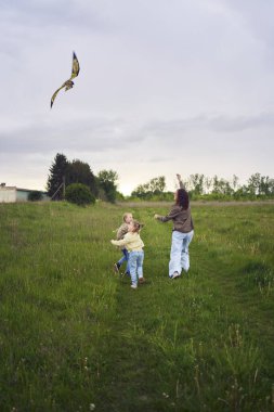 İki küçük kız kardeş ve anne koşup tarlaya uçurtma fırlatıyorlar.