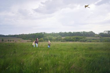 İki küçük kız kardeş ve anne koşup tarlaya uçurtma fırlatıyorlar.