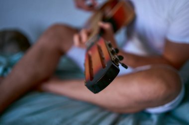 Genç adam evde gitar çalmayı öğreniyor, yanında bir kedi yatıyor ve dinliyor.