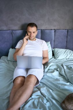 Yatakta dizüstü bilgisayarda çalışan ve telefonla konuşan genç adam.                         