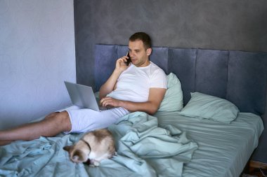 Yatakta dizüstü bilgisayarda çalışan ve telefonla konuşan genç adam.                         