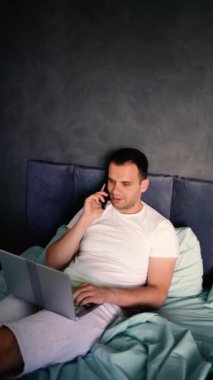 Yatakta dizüstü bilgisayarda çalışan ve telefonla konuşan genç adam.
