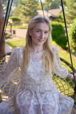 Pastel şifon elbiseli genç sarışın bir kadının portresi hasır bir hamakta daireler çiziyor.