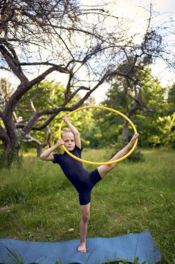 5 yaşındaki küçük bir jimnastikçi. Siyah, minimalist bir mayo giymiş. Bahçede bir halkayla egzersiz yapıyor.
