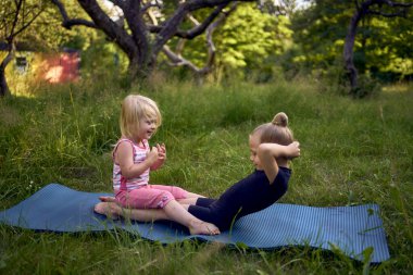 Siyah minimalist tulumlu 5 yaşındaki jimnastikçi, yürümeye yeni başlayan kız kardeşinin egzersizlerini gösteriyor.