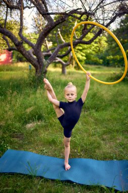 5 yaşındaki küçük bir jimnastikçi. Siyah, minimalist bir mayo giymiş. Bahçede bir halkayla egzersiz yapıyor.