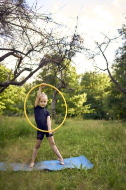 5 yaşındaki küçük bir jimnastikçi. Siyah, minimalist bir mayo giymiş. Bahçede bir halkayla egzersiz yapıyor.