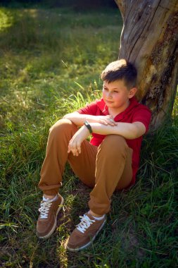 Kırmızı tişörtlü bir çocuk parktaki solmuş bir ağacın yanında üzgün.