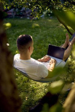 Adamın biri evcil köpeğinin yanındaki hamakta dizüstü bilgisayarda çalışıyor.