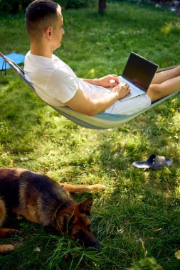 Adamın biri evcil köpeğinin yanındaki hamakta dizüstü bilgisayarda çalışıyor.