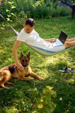 Adamın biri evcil köpeğinin yanındaki hamakta dizüstü bilgisayarda çalışıyor.