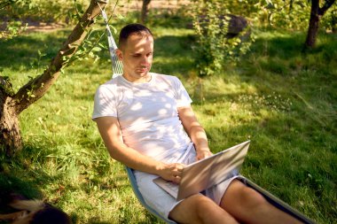 Adamın biri evcil köpeğinin yanındaki hamakta dizüstü bilgisayarda çalışıyor.