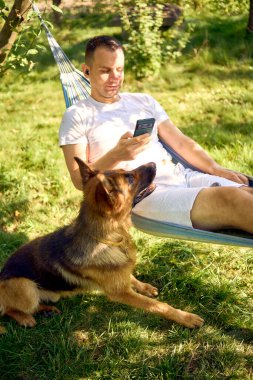 Bir adam dinlenir ve hamakta müzik dinler, köpeği yakınlarda dinlenir.