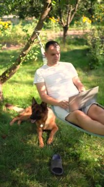 Adamın biri evcil köpeğinin yanındaki hamakta dizüstü bilgisayarda çalışıyor.
