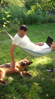 Adamın biri evcil köpeğinin yanındaki hamakta dizüstü bilgisayarda çalışıyor.