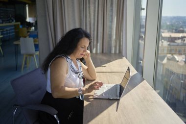 57 yaşındaki bir kadın, modern bir ofis çalışma alanında dizüstü bilgisayarda çalışıyor.                    