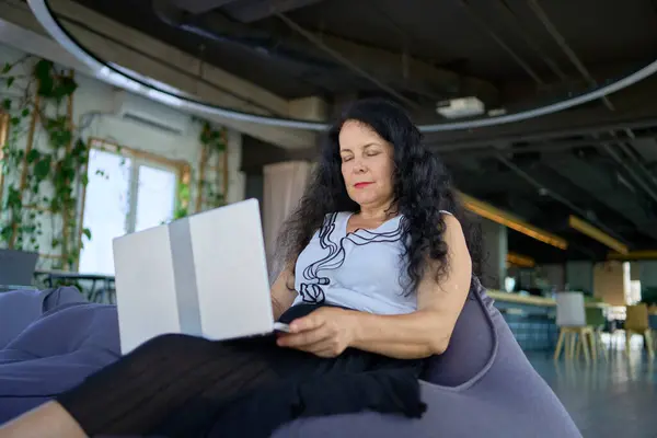 stock image a  57-year-old woman works on a laptop in a modern office workspace  sitting in a beanbag chair                  