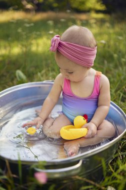 Metal bir küvette bir bebek yaz bahçesinde plastik ördek yavrularıyla oynuyor.