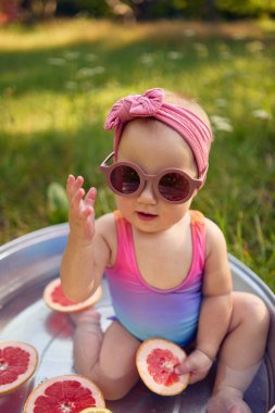 Bir yaz bahçesinde metal bir banyoda güneş gözlüğü takan bir bebek greyfurt ve limon dilimleriyle oynar.