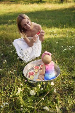 Metal bir banyoda, yaz bahçesinde anne ve kız kardeşin greyfurt yemesini izleyen bir bebek.                       