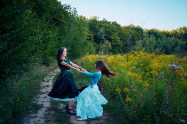 two woman in a whimsical aesthetic style dress with a german shepherd in a field in the summer at a sunset clipart