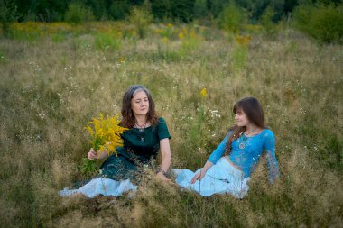 two woman in a whimsical aesthetic style dress with a german shepherd in a field in the summer at a sunset clipart