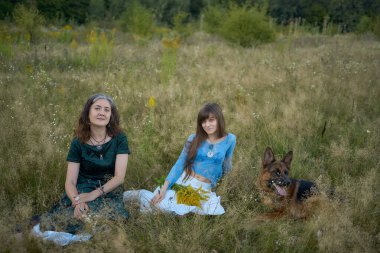 two woman in a whimsical aesthetic style dress with a german shepherd in a field in the summer at a sunset clipart