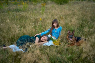 two woman in a whimsical aesthetic style dress with a german shepherd in a field in the summer at a sunset clipart