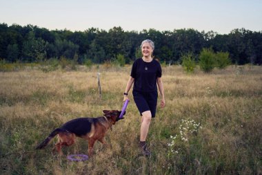 İki kadın gün batımında bir Alman çoban köpeği oyuncağıyla sahada oynuyor.