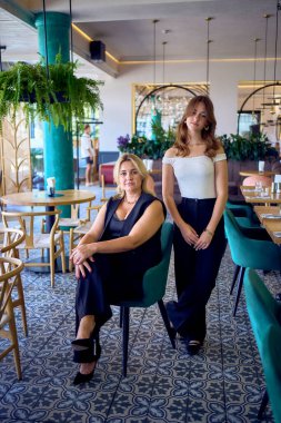           Orta yaşlı, iri yarı bir kadın ve genç kızı lüks bir restoranda eski moda kıyafetler giyiyorlar.                                        