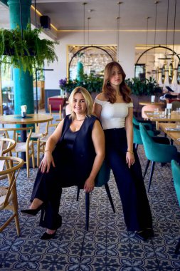           Orta yaşlı, iri yarı bir kadın ve genç kızı lüks bir restoranda eski moda kıyafetler giyiyorlar.                                        